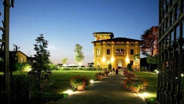 A large, illuminated building with a central tower, surrounded by a well-maintained garden and a paved pathway leading to the entrance. - Villa Moorings (Barga)