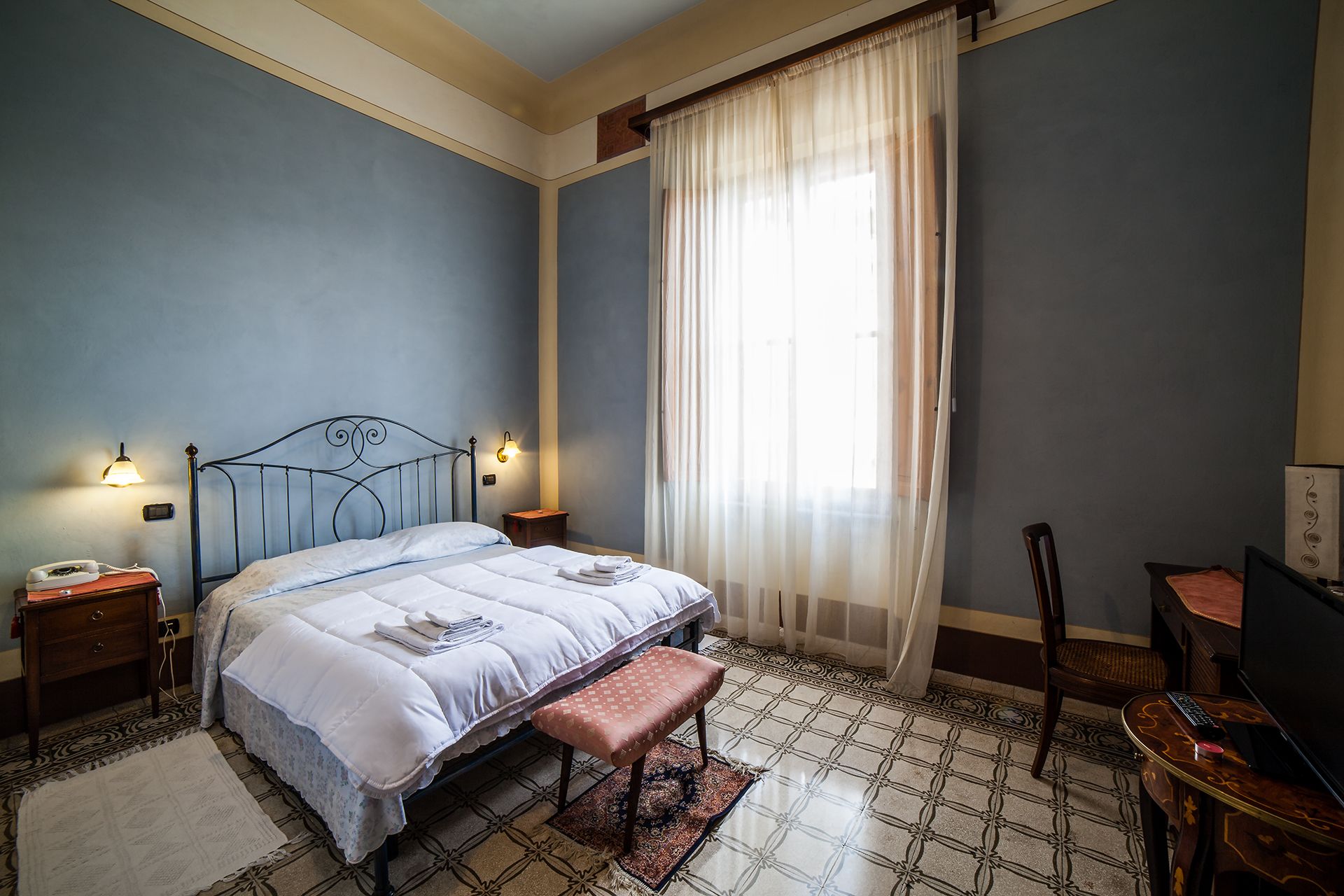 Une chambre à coucher avec un lit, des lampes de chevet, une chaise, un bureau et une fenêtre avec des rideaux légers.
