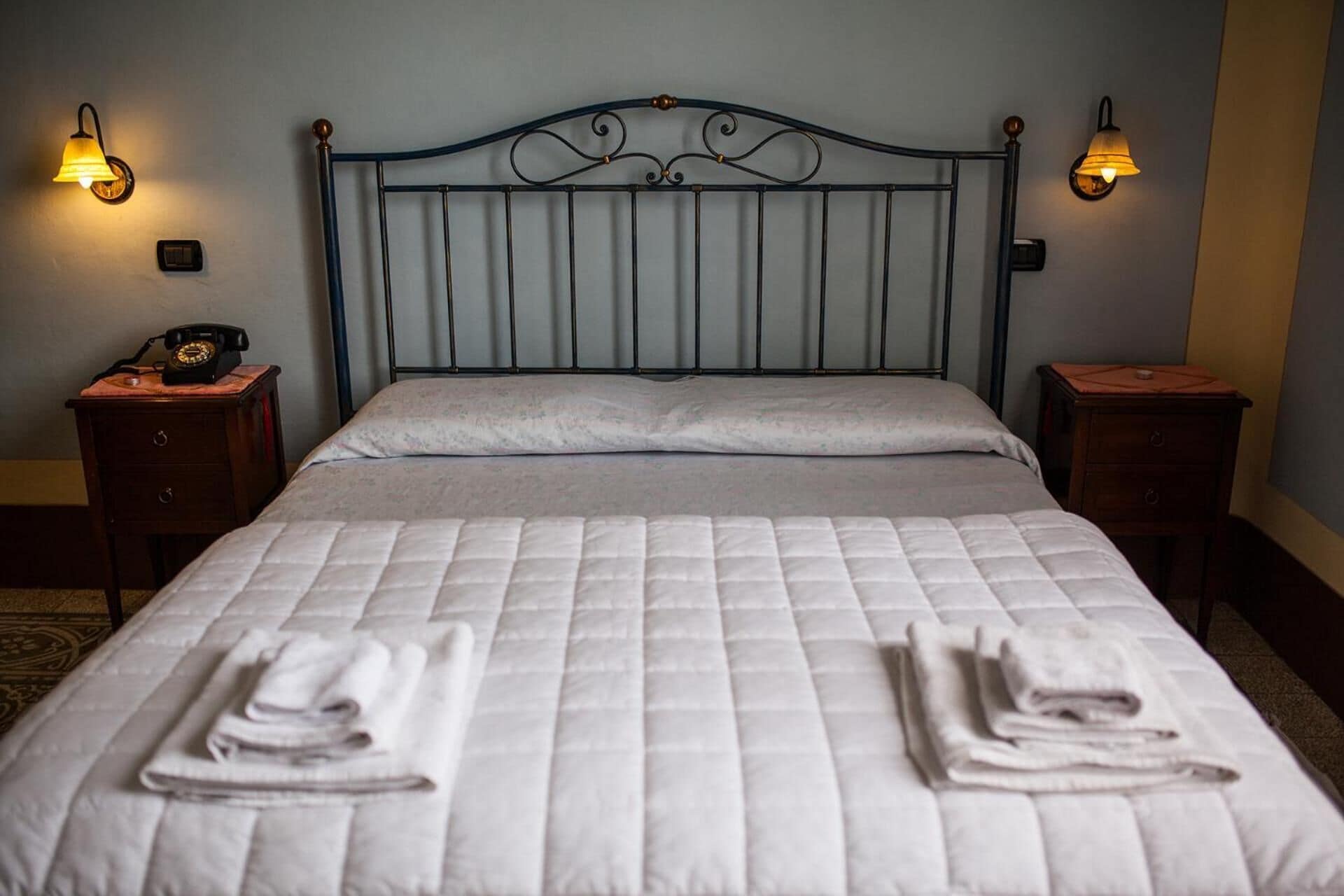 Un letto rifatto con un piumone bianco trapuntato, due asciugamani piegati sul letto e due comodini con lampade su entrambi i lati.