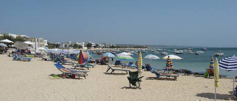 A beach with rows of sun loungers and umbrellas, a clear sky, and boats in the water.