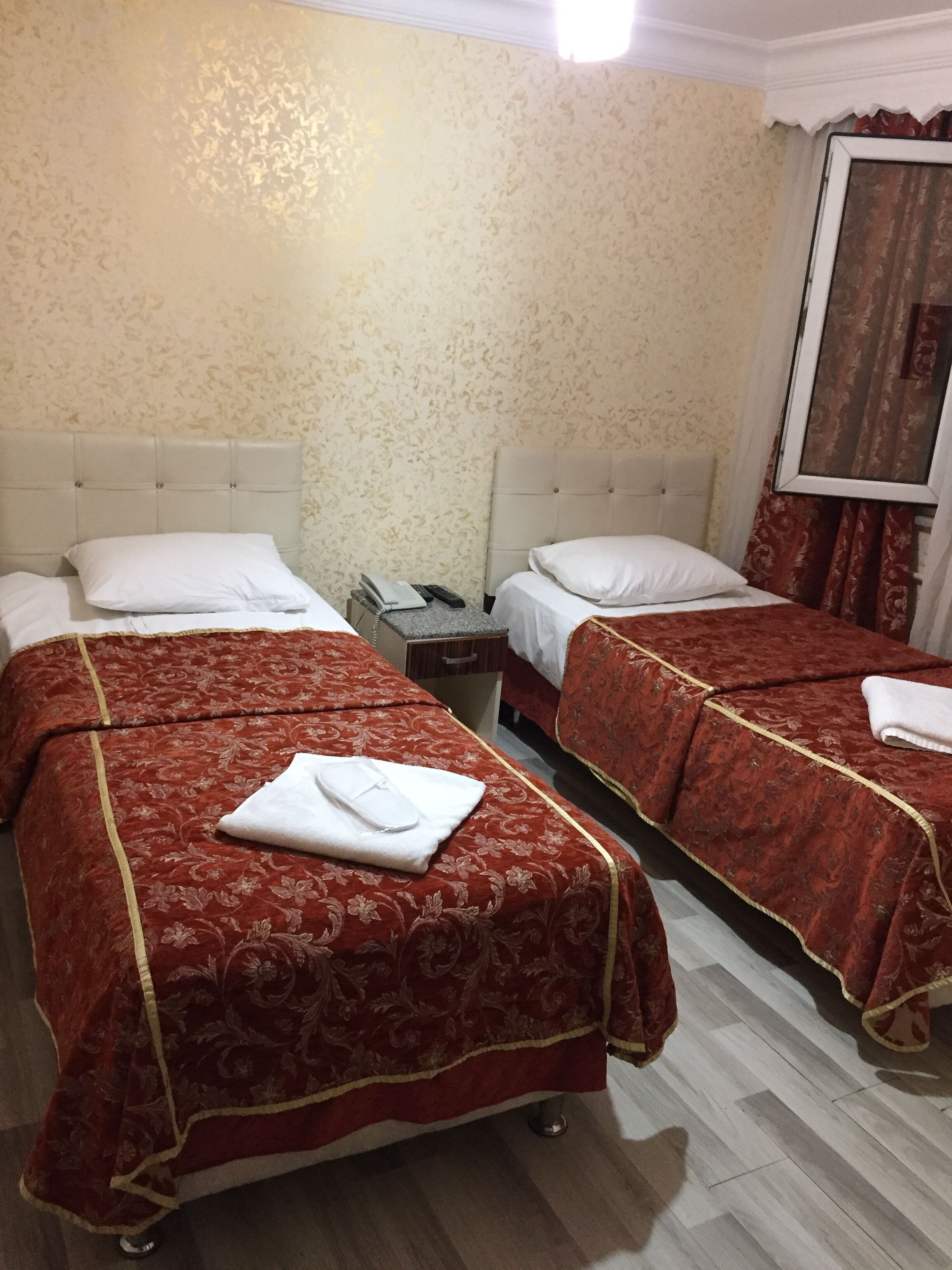 A hotel room with two beds, each with a red patterned bedspread and white pillows. There is a small bedside table with a telephone and a lamp. The room has a light-colored wall and a window with curtains.