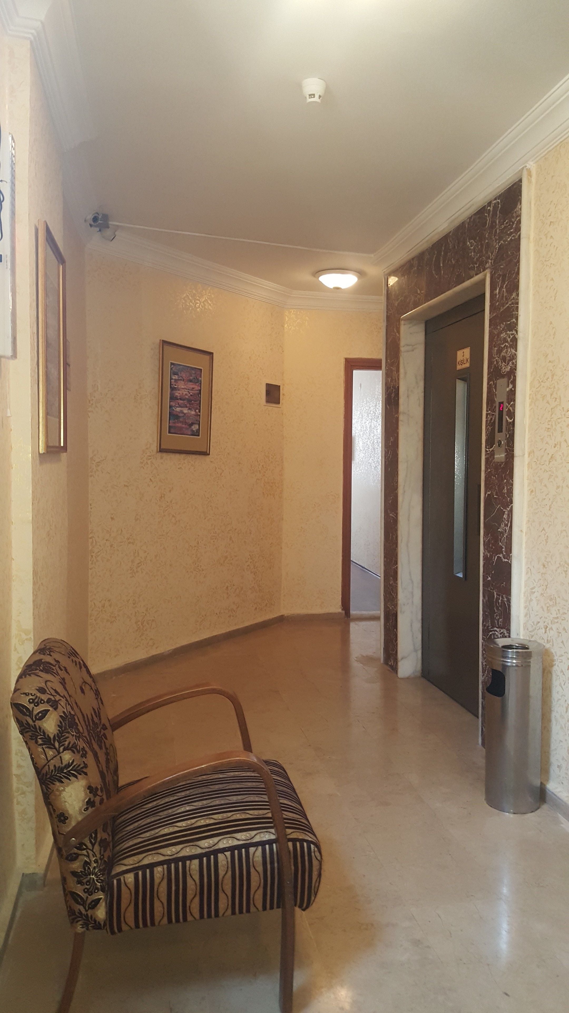 A hallway with a patterned chair, a trash can, and a door with a sign.