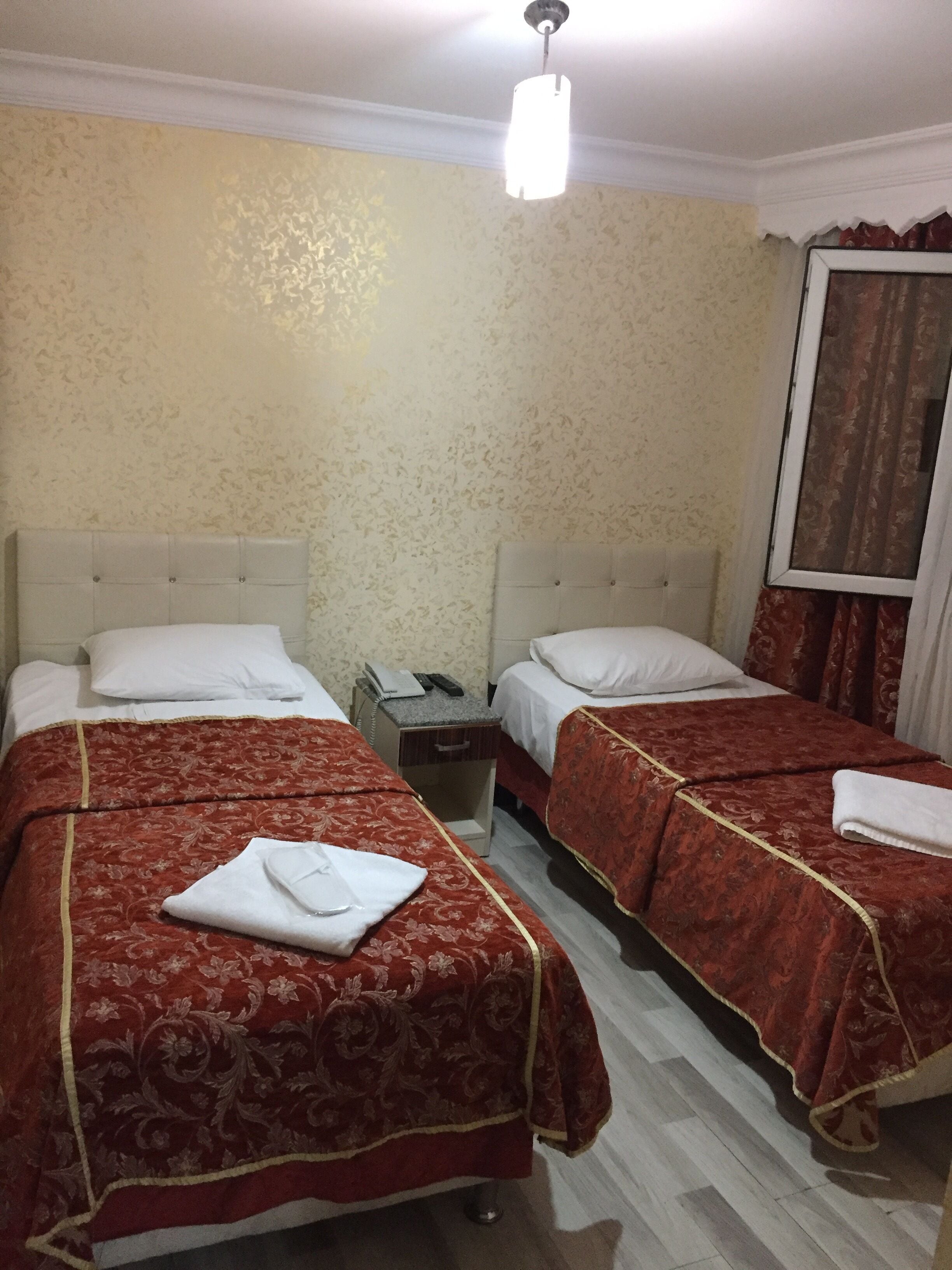 A hotel room with two beds, each with a red bedspread and white pillows. There is a nightstand between the beds with a telephone and a lamp. The walls have a textured wallpaper, and there is a window with curtains.