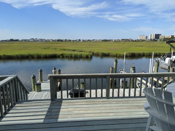 A wooden deck with a railing overlooking a calm body of water, a grassy field, and distant buildings. - Experience Ocean City like never before at Sea Grass #5! (Ocean City)