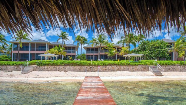 A wooden pier leading to a beachfront hotel with multiple balconies and palm trees. - Northern Lights #5: 2-Story Townhome w/Backyard Snorkeling, Shared Pool + Dock (Old Man Bay)