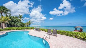 A swimming pool with lounge chairs and a thatched structure, surrounded by greenery and a view of the ocean. - Northern Lights #5: 2-Story Townhome w/Backyard Snorkeling, Shared Pool + Dock (Old Man Bay)