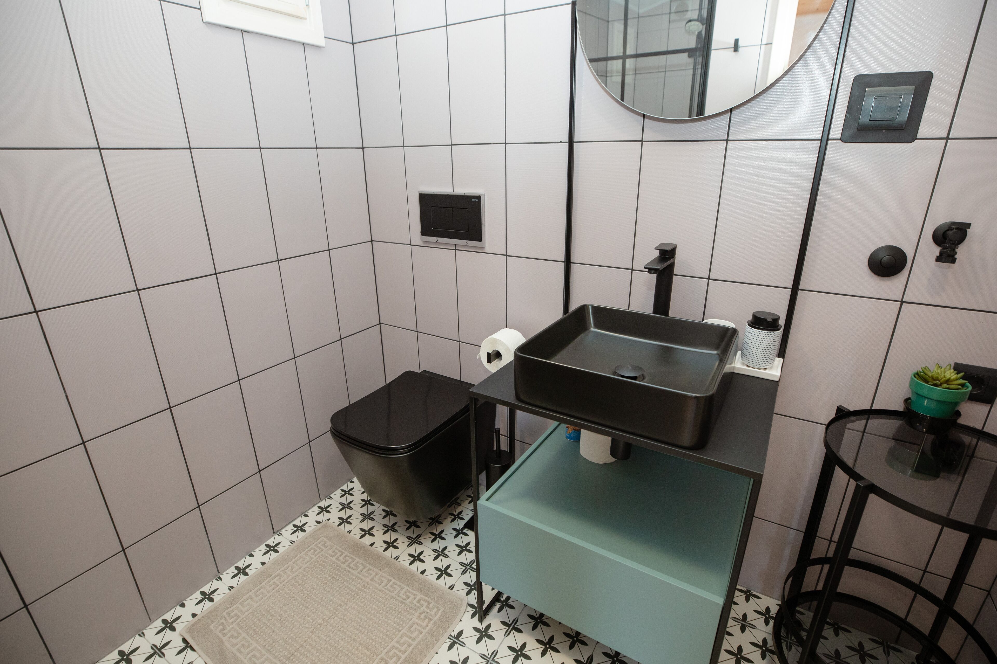 Een badkamer met een zwarte prullenbak, een toiletrolhouder, een wastafel met spiegel en een kleine groene plant op een tafeltje.