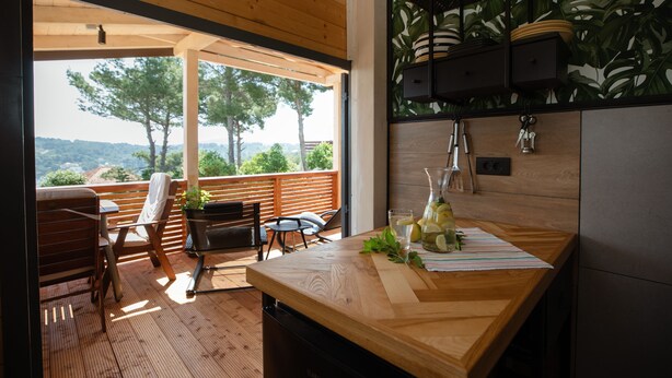 A wooden deck with outdoor seating, a kitchen area with a wooden countertop, and a view of trees and hills.