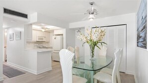 A dining area with a glass table surrounded by white chairs, a white kitchen in the background, and a ceiling fan. - Beachfront 2BR/2BA condo with pool, wifi, and Marco Island views. (Marco Island)