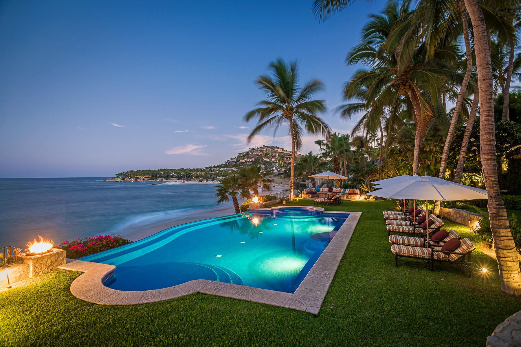 A swimming pool with a curved edge, surrounded by grass and palm trees, with a view of the ocean and a hillside in the background.