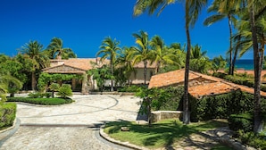Un resort tropical con un camino pavimentado que conduce a un edificio de techo con tejas, rodeado de palmeras y un cielo azul despejado.