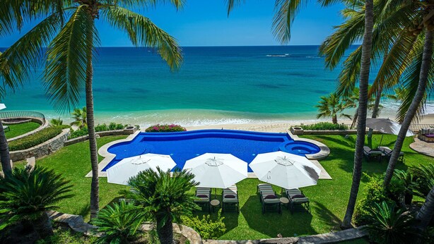Una piscina con sillones de descanso y sombrillas, rodeada de palmeras y vista al océano.