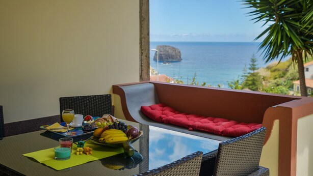 Un balcon avec une table préparée pour le petit-déjeuner, comprenant des fruits et des boissons, et offrant une vue sur la côte.
