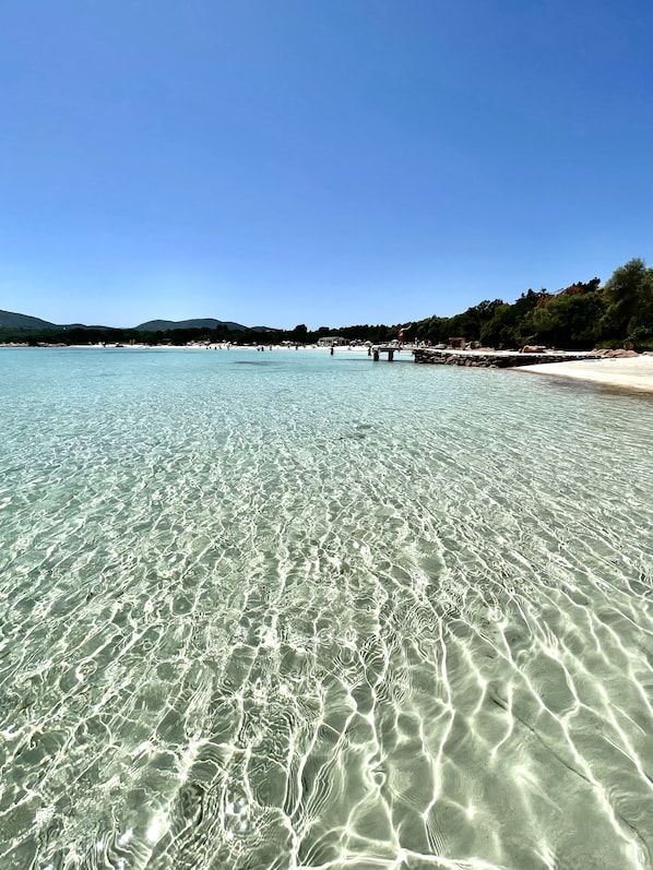 A clear beach with shallow water, a sandy shore, and a distant view of hills.