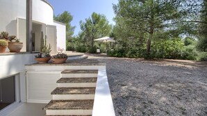 Un bâtiment moderne à l’extérieur blanc, avec une allée en gravier et un escalier en pierre menant à un espace extérieur couvert, équipé d’une table et de chaises.