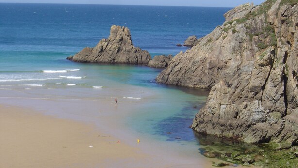 A beach with clear waters, rocky cliffs, and a person walking on the shore.