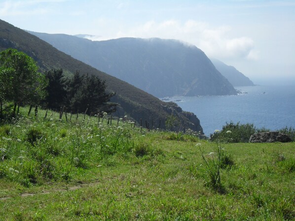 Paisaje costero con un primer plano de hierba, una valla de madera y vistas al mar y a las montañas al fondo.