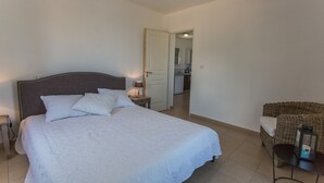 Un dormitorio con una cama grande, una mesita de noche con lámpara, una silla de mimbre y vistas a la cocina.