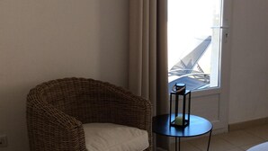 Una habitación con una silla de mimbre, una mesita redonda pequeña, una cama, una ventana con cortinas y un cuadro enmarcado en la pared.