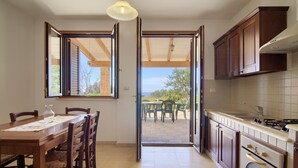 A kitchen with wooden cabinets, a dining table with chairs, and a view through an open door to an outdoor patio. - Il Cedro San Gregorio by LoveSud (Marina San Gregorio)