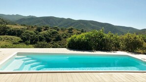A rectangular swimming pool with clear blue water, surrounded by a wooden deck. In the background, there are green hills and a clear sky. - Refined independent villa for design lovers in the beautiful Chia bay (Domus De Maria - Chia)