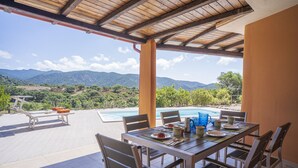 A covered outdoor dining area with a pool, chairs, and a table set for a meal, overlooking a mountainous landscape. - Refined independent villa for design lovers in the beautiful Chia bay (Domus De Maria - Chia)