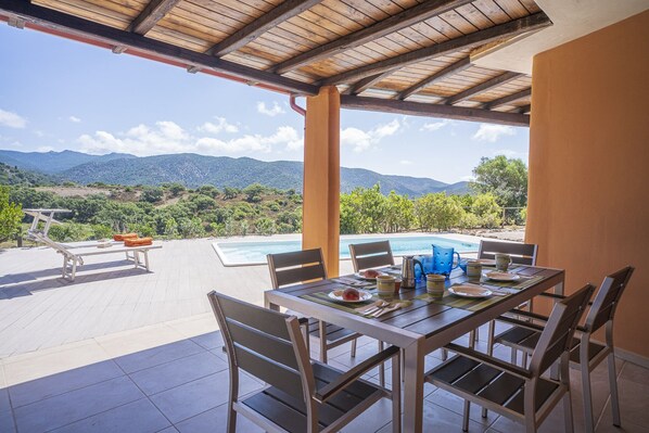 A covered outdoor dining area with a pool, chairs, and a table set for a meal, overlooking a mountainous landscape.