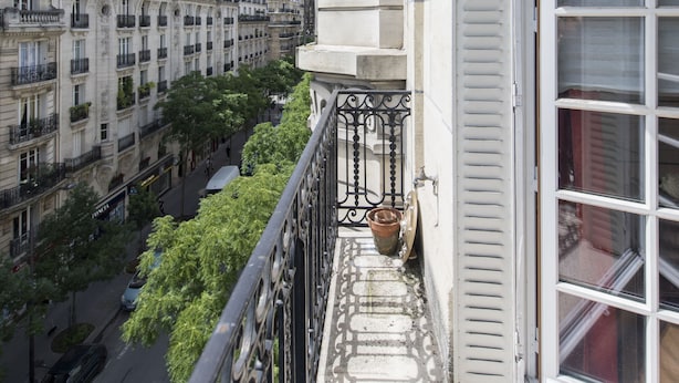 Een balkon met decoratieve hekwerk, een bloempot en uitzicht op een straat met gebouwen en bomen.