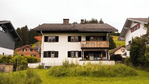 Uma casa de dois andares com varanda, cercada por vegetação e outras residências.