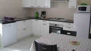 Cocina compacta con armarios blancos, estufa a gas, microondas y una mesa con mantel estampado de cerezas.