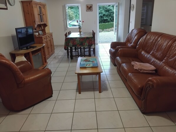 A living room with a brown leather sofa, a wooden coffee table, a television on a stand, and a dining table with chairs.