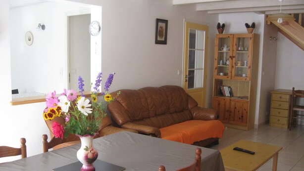 Una sala de estar con un sofá de cuero marrón, una mesa de comedor con sillas, un aparador de madera y un florero con flores.