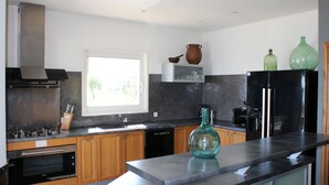 A modern kitchen with wooden cabinets, a stainless steel range hood, a built-in oven, a black dishwasher, and a large window providing natural light. - Contemporary villa, with clim-160 m2, swimming pool, 5 mn from beaches & shops (Monticello)