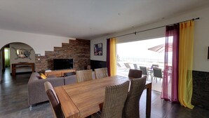 A modern living room with a wooden dining table, wicker chairs, a flat-screen TV, and a stone wall. - Contemporary villa, with clim-160 m2, swimming pool, 5 mn from beaches & shops (Monticello)