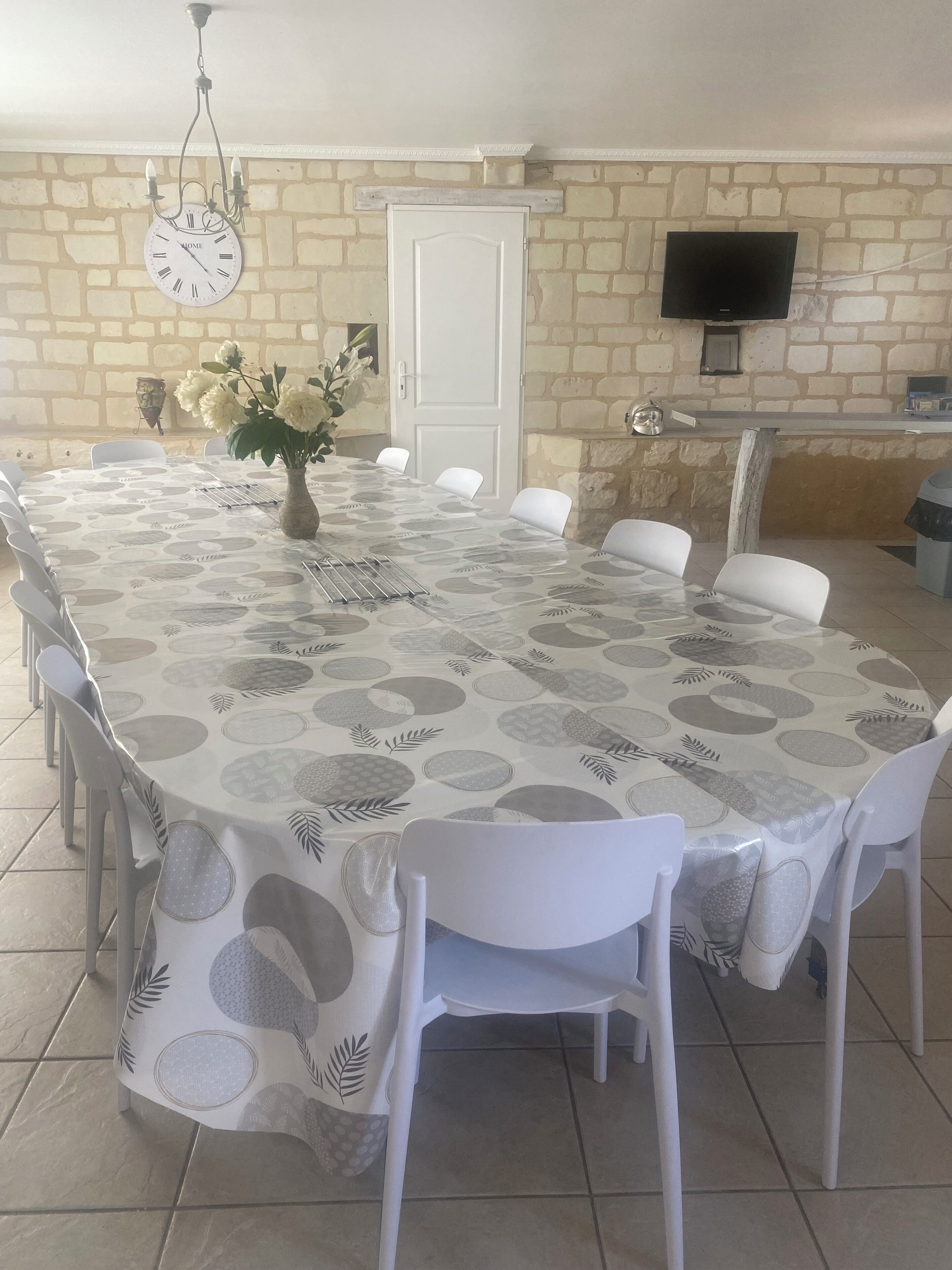 A dining area with a large table covered in a leaf-patterned tablecloth, surrounded by white chairs. A wall-mounted clock and a television are visible in the background.