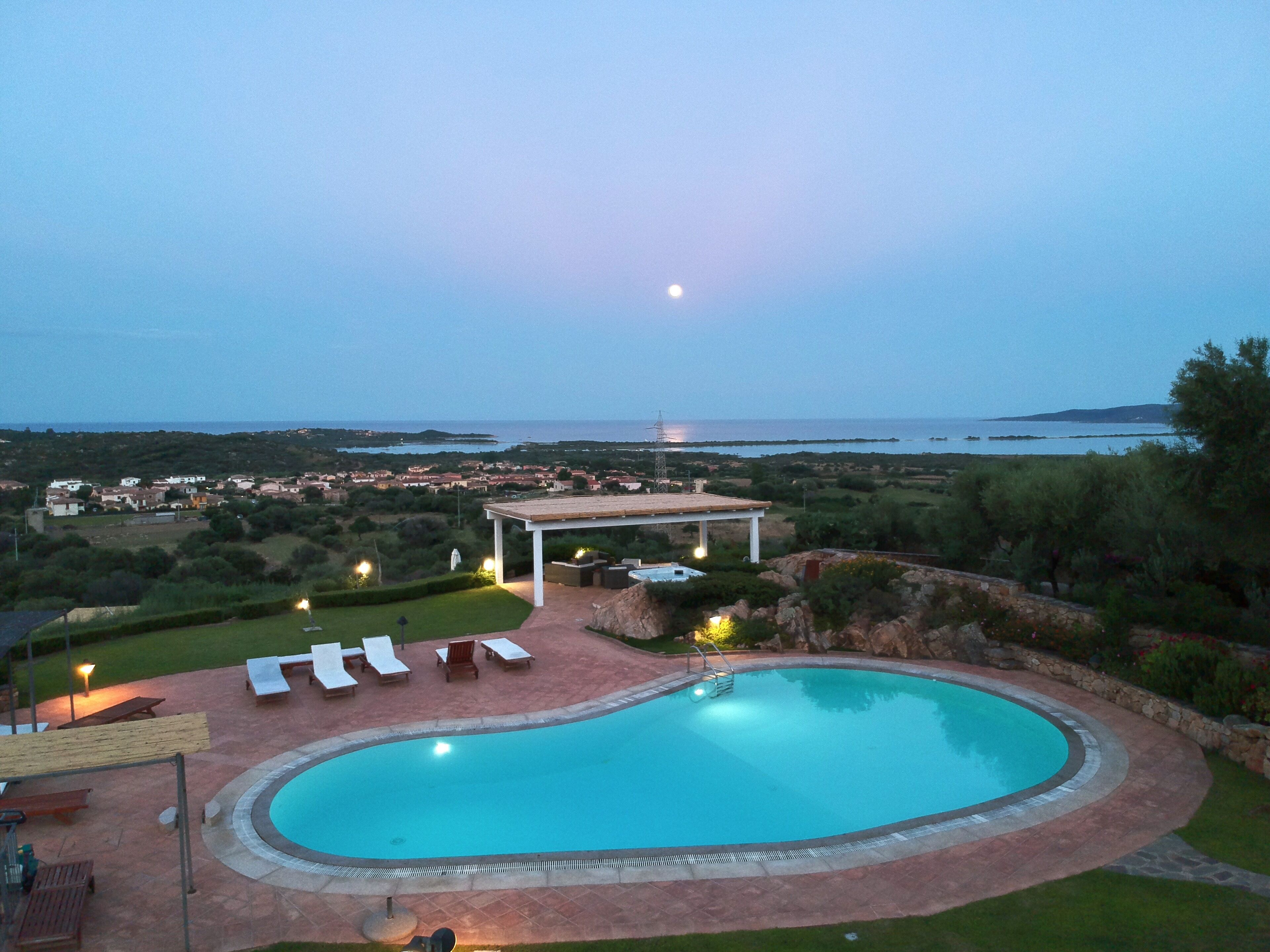 Une piscine avec des chaises longues, une pergola et une vue sur la mer et la ville au crépuscule.