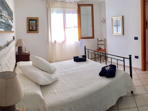 Une chambre à coucher avec un lit, des oreillers, une chaise, une lampe, une fenêtre avec des rideaux et un tableau au mur.