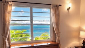 A bedroom with a large window offering a view of the ocean and greenery, a wooden dresser, a bed with white bedding, and a wall-mounted lamp.