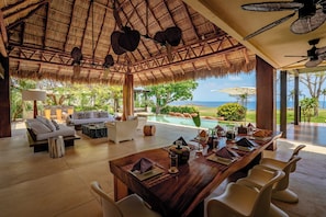 A spacious indoor area with a dining table set for six, a sofa, and a pool visible through the open space.