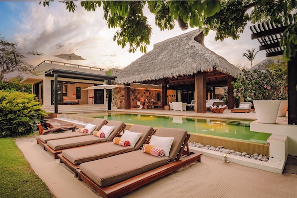 A poolside area with lounge chairs, a thatched-roof structure, and a pool.