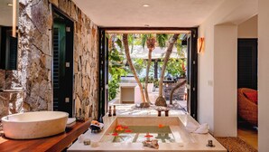 Un área de spa con pared de piedra, un lavamanos grande, una bañera de hidromasaje y vistas a un jardín tropical.