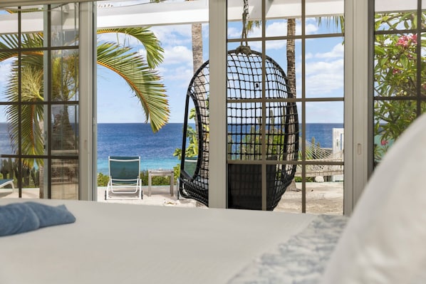 A room with a bed, a hanging chair, and a view of the ocean through large windows.