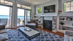 A spacious living room with a fireplace, a television mounted above it, and a view of the ocean through large sliding doors. - AMAZING Lakefront And Well Designed Lakehouse: That Is Sure To Please (Romulus)