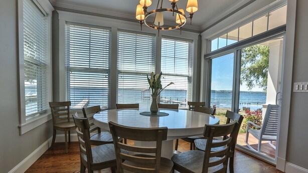 A dining area with a round table and chairs, large windows with blinds, and a view of the outdoors. - AMAZING Lakefront And Well Designed Lakehouse: That Is Sure To Please (Romulus)