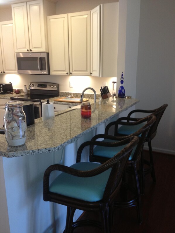 A kitchen with white cabinets, a microwave, an oven, a granite countertop, and a bar area with four chairs.