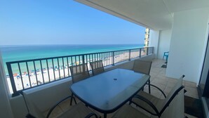 Un balcón con una mesa y sillas de vidrio, con vista a una playa con sombrillas y un cielo azul despejado.