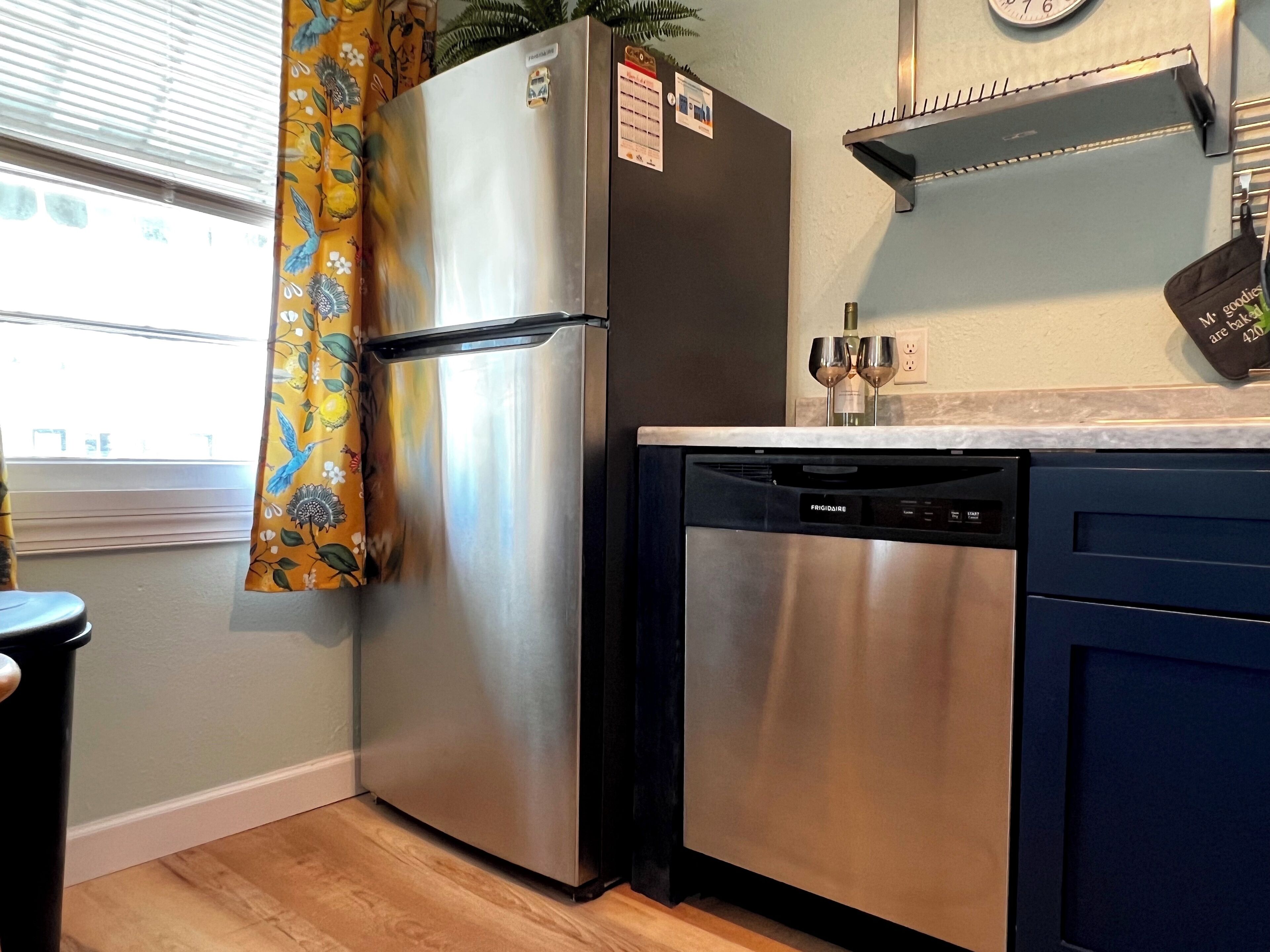 A kitchen with a stainless steel refrigerator, a dishwasher, and a window with curtains.