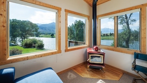 Una habitación con ventanas amplias que dan a un lago y montañas, techo de madera y estufa antigua.