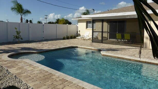 A swimming pool with a curved design, surrounded by a paved patio and a white fence.