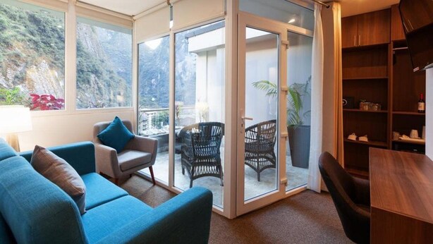 A modern living room with a blue sofa, a small armchair, and a dining table. There is a sliding glass door leading to an outdoor area with wicker chairs and a potted plant. - Inti Punku Alameda Inn (Machu Picchu)